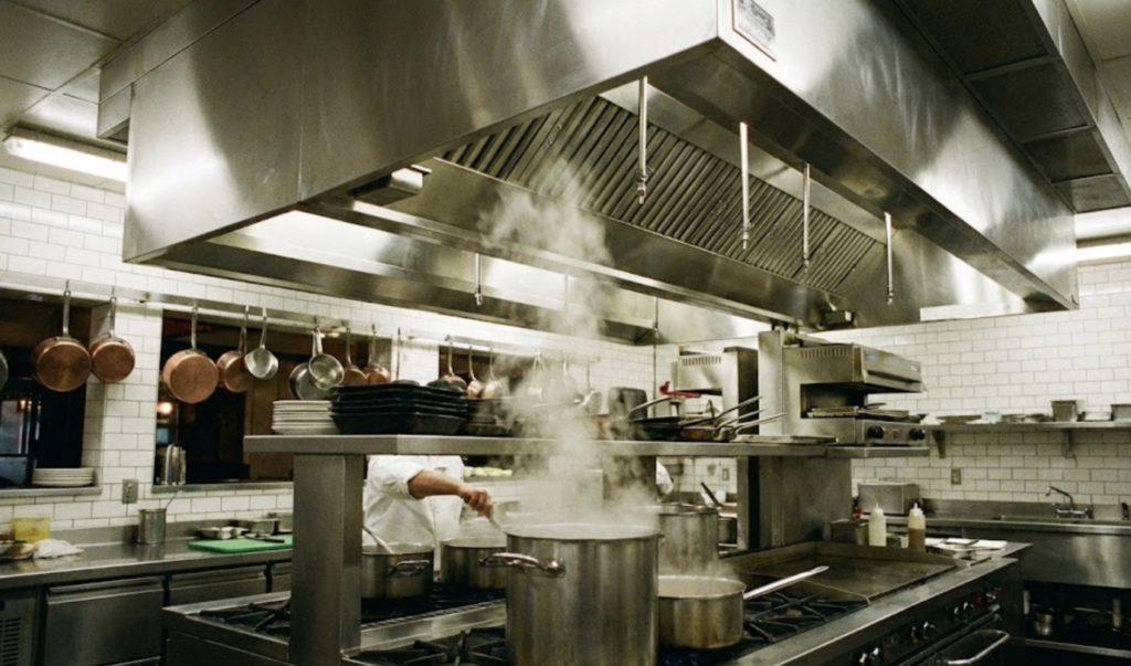 Grande hotte en inox dans une cuisine de restaurant en pleine activité, illustrant l'importance du nettoyage de hotte professionnelle.
